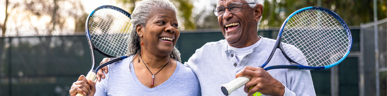 An elderly senior living couple standing on a tennis court holding tennis rackets while smiling