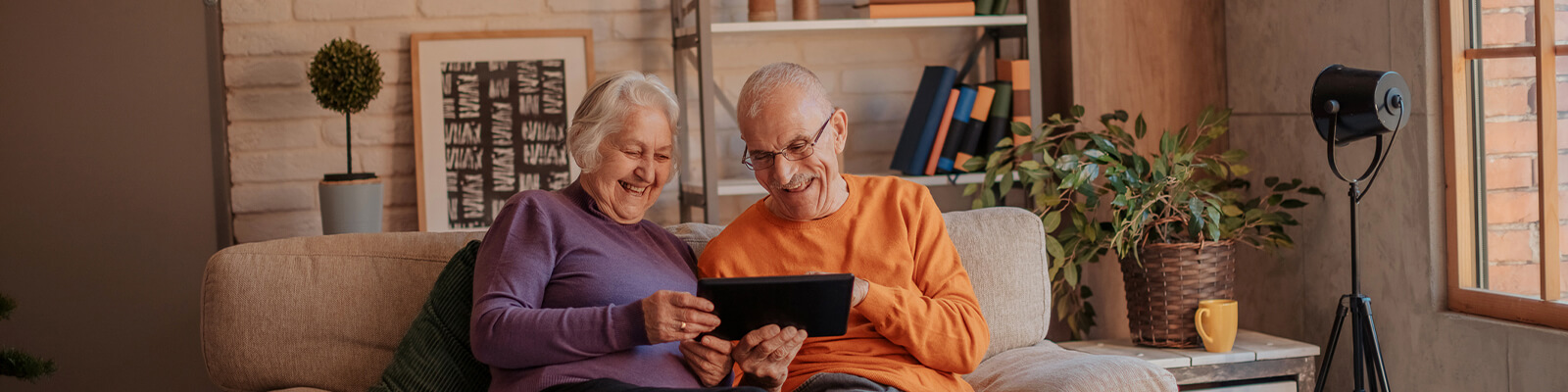 Elderly man and woman sitting on a tan couch smiling and looking at a smart tablet