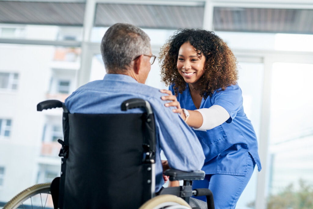 A female senior nursing assistant in blue scrubs leaning down to talk to a male senior living resident sitting in a wheelchair